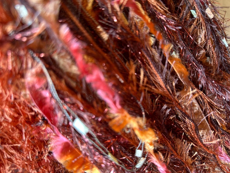 May include: Close-up of richly textured yarn fringe  in shades of deep red, burnt orange, and copper.  The fibers are long and slightly fuzzy, creating a sense of depth and movement.  Metallic threads are subtly interwoven, adding shimmer and visual interest.  They add a luxurious, multi-toned effect.