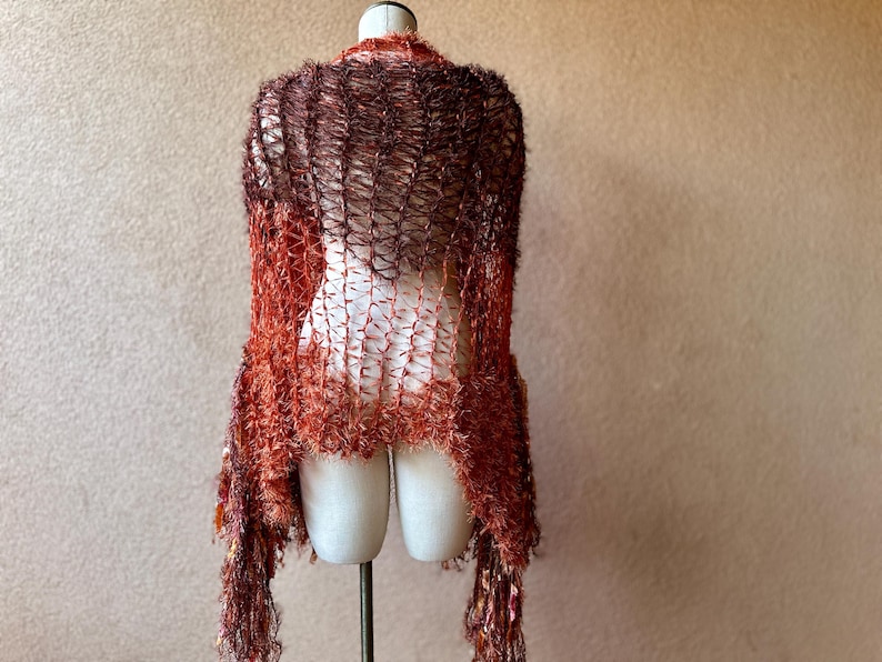 May include: A brown and orange crochet shawl with a fringe border. The shawl is draped over a mannequin.
