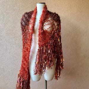 May include: A red and brown crocheted shawl with fringe. The shawl is displayed on a white mannequin.