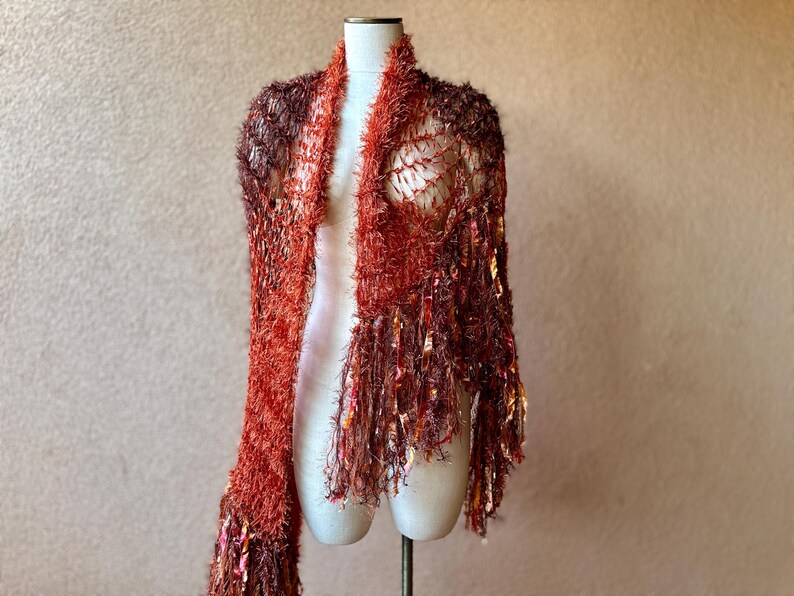 May include: A red and brown crocheted shawl with fringe. The shawl is displayed on a white mannequin.