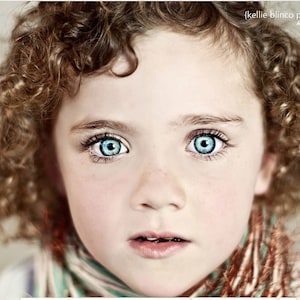 May include: A young girl with curly brown hair and bright blue eyes. She is wearing a scarf with a brown, white, and green pattern.