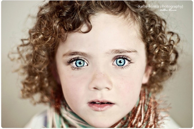 May include: A young girl with curly brown hair and bright blue eyes. She is wearing a scarf with a brown, white, and green pattern.