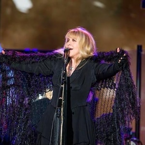 May include: A woman with blonde hair sings into a microphone while wearing a black dress and a black shawl with a fringe. The shawl is a dark purple color. The woman is on a stage with a microphone stand in front of her.