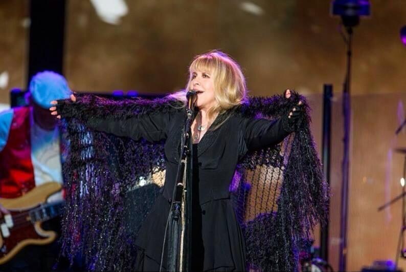 May include: A woman with blonde hair sings into a microphone while wearing a black dress and a black shawl with a fringe. The shawl is a dark purple color. The woman is on a stage with a microphone stand in front of her.