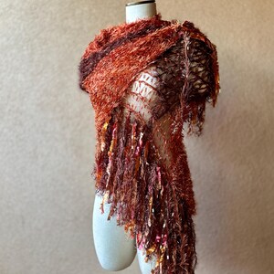 May include: A rust-colored fuzzy scarf with a crocheted design and long fringe. The scarf is draped over a white mannequin.