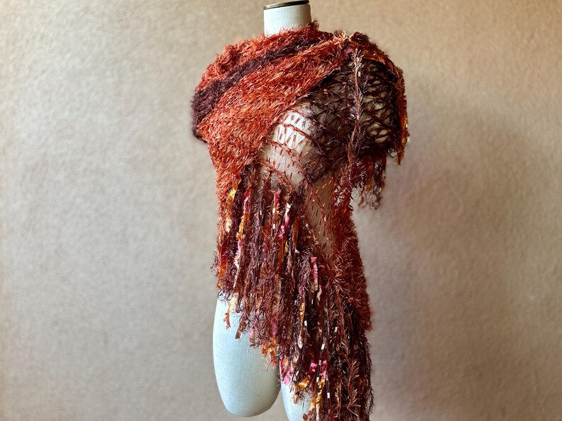 May include: A rust-colored fuzzy scarf with a crocheted design and long fringe. The scarf is draped over a white mannequin.