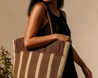 Puede incluir: Bolso tote tejido con rayas verticales marrones y crema. El borde superior presenta flecos marrones y el bolso tiene asas marrones. El bolso lo lleva una persona que viste un vestido negro.