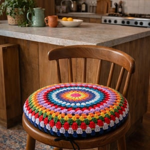 Op de afbeelding: Een ronde, kleurrijke gehaakte zitkussen in regenboogkleuren, waaronder rood, oranje, geel, groen, blauw en paars. Het kussen ligt op een houten stoel. De achtergrond toont een keuken met een aanrecht en apparaten.