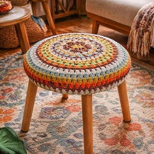 Könnte beinhalten: Ein runder, niedriger Hocker mit einer farbenfrohen, gehäkelten Oberseite. Die Oberseite weist konzentrische Kreise in Braun, Gelb, Orange, Grün und Blau auf. Der Hocker hat vier Holzbeine. Der Hocker steht auf einem gemusterten Teppich.
