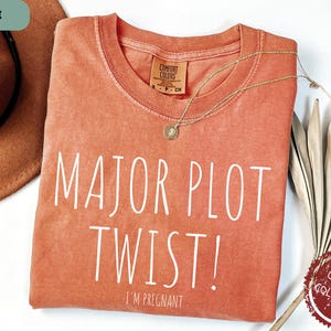May include: A peach-coloured t-shirt with the text "MAJOR PLOT TWIST! I'M PREGNANT" in white. The shirt is made of a soft, comfortable fabric.