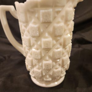 Puede incluir: Jarra blanca de estilo vintage con un patrón de diamantes y un borde festoneado. La jarra tiene un asa y un pico para verter. El vidrio es opaco y tiene una superficie texturizada. Esta jarra decorativa es perfecta para servir bebidas.