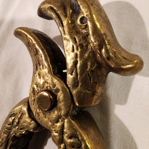 May include: Close-up of a vintage brass nutcracker. The nutcracker is shaped like a bird with detailed features, including textured feathers and a beak-like opening. The brass has a warm, aged patina, suggesting its vintage nature.