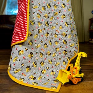 May include: A gray baby quilt with a pattern of yellow construction vehicles and road signs. The quilt has a red and white checkered backing and a yellow border. A yellow and orange toy excavator sits on the quilt.