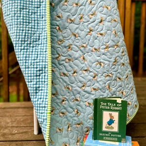May include: A light blue baby quilt with a repeating pattern of rabbits, paired with a stack of children's books, including "The Tale of Peter Rabbit" by Beatrix Potter. The quilt has a green and white gingham backing and trim.