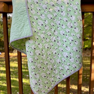 May include: A light green baby quilt with a pattern of white sheep with black faces. The quilt has a light green backing with a quilted pattern and a gray and white striped border. The quilt is draped over a wooden railing.