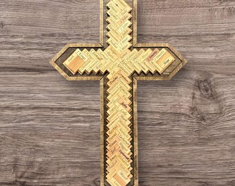 May include: Wooden cross with a layered design. The cross features the names of Bible books in a decorative pattern. The cross is made of light brown wood and is displayed against a wood grain background. The cross is a religious item.