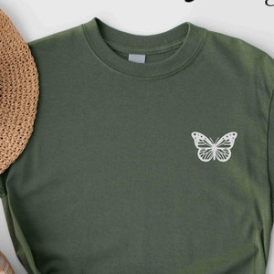 May include: Military green t-shirt with a white butterfly graphic. The shirt is knotted at the bottom and paired with a straw hat and patterned shoes. The words "Military Green" are written in cursive at the top.