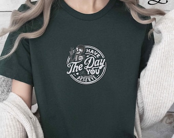 May include: Forest green t-shirt with a round white graphic that reads "Have The Day You Deserve" with a skeleton and hand illustration. The shirt is a solid color and has short sleeves.