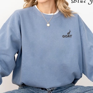 May include: A light blue sweatshirt with a black goat graphic and the word "GOAT" printed below it. The sweatshirt is worn by a person wearing blue jeans.