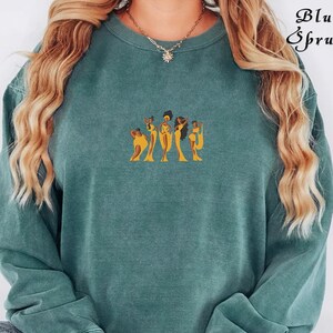 Op de afbeelding: Een tealgroen sweatshirt met een afbeelding van vijf vrouwen in gele jurken. De vrouwen staan in een rij en hebben een donkere huid en haar.