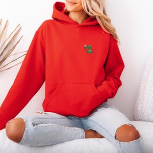 May include: A bright red hoodie with a kangaroo pocket and a drawstring hood. The hoodie features a small embroidered design of a green frog with a red heart on the chest. The model is wearing light blue ripped jeans.