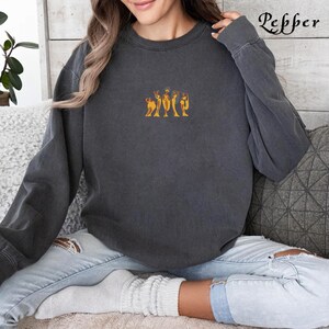 Op de afbeelding: Een donkergrijs sweatshirt met een grafische afbeelding van vijf figuren in een rij. De figuren zijn oranje en zwart en lijken gewaden te dragen. Het sweatshirt wordt gedragen door een persoon die op een bank zit.