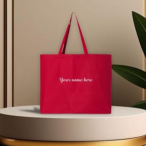 May include: A red tote bag with long handles, featuring the text "Your name here" in a cursive font. The bag is displayed on a white pedestal with a gold trim, against a neutral background. The bag is made of a sturdy material.