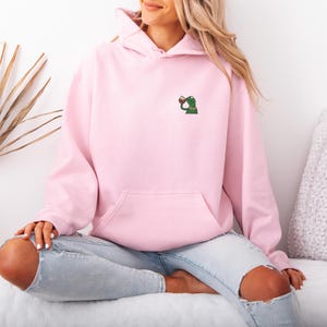 May include: A light pink hoodie with a kangaroo pocket and a small embroidered frog design on the chest. The frog is holding a cup. The hoodie is worn with light blue jeans.