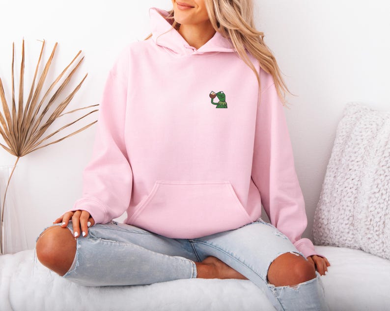 May include: A light pink hoodie with a kangaroo pocket and a small embroidered frog design on the chest. The frog is holding a cup. The hoodie is worn with light blue jeans.