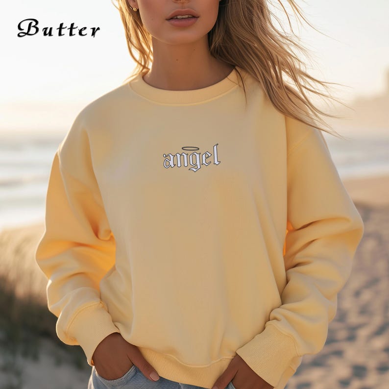 Pode incluir: Uma camisa de moletom amarelo claro com um design bordado branco que diz "angel" com uma aur&eacute;ola acima da palavra.