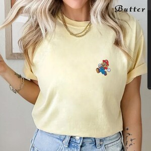 May include: A yellow short-sleeved t-shirt with a cartoon image of Mario, a video game character, on the front. The character is wearing a red hat and blue overalls.