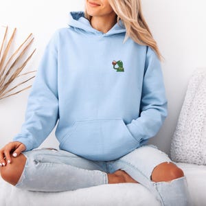 May include: Light blue hoodie with a kangaroo pocket and a drawstring hood. The hoodie features a small embroidered design of a green frog drinking from a red cup on the upper left chest. The model is wearing light blue jeans.