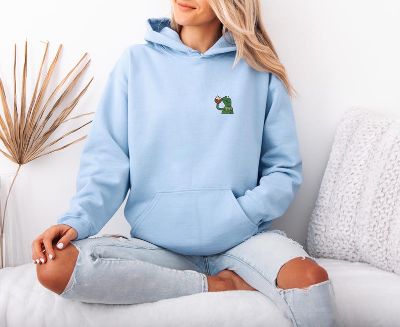 May include: Light blue hoodie with a kangaroo pocket and a drawstring hood. The hoodie features a small embroidered design of a green frog drinking from a red cup on the upper left chest. The model is wearing light blue jeans.