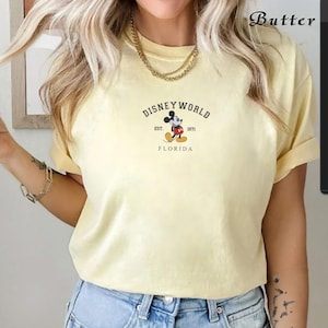 Puede incluir: Camiseta amarilla clara con el logotipo de Disney World y Mickey Mouse. El logotipo dice "Disney World", "Est. 1971" y "Florida". La camiseta tiene mangas cortas y cuello redondo.