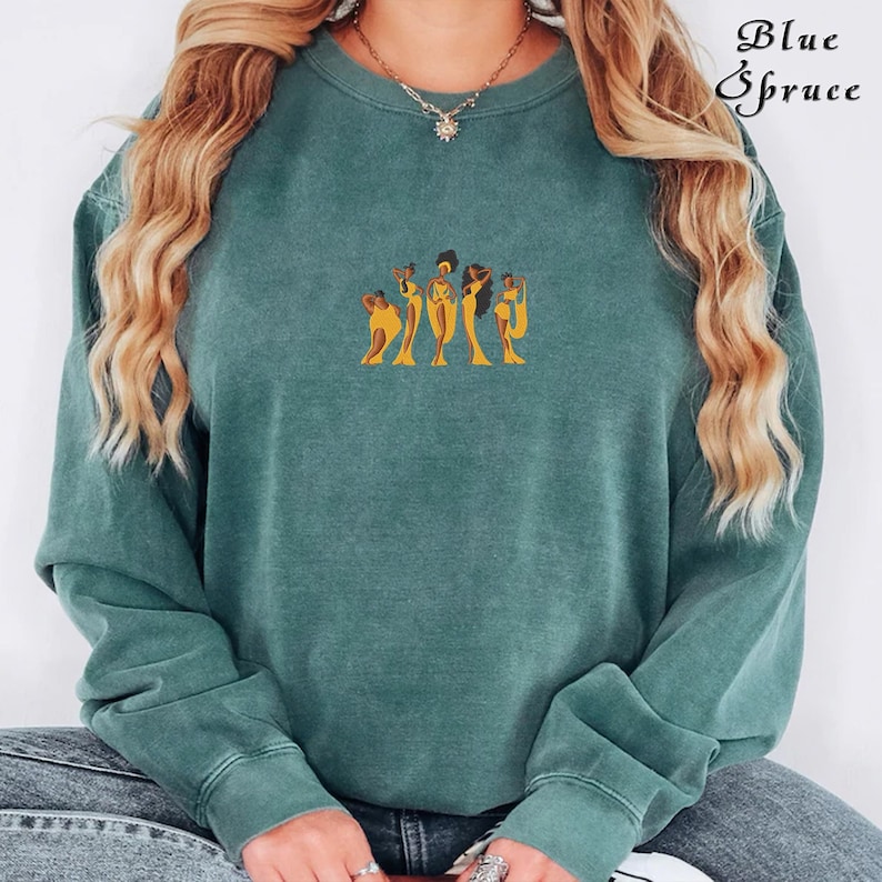 Op de afbeelding: Een tealgroen sweatshirt met een afbeelding van vijf vrouwen in gele jurken. De vrouwen staan in een rij en hebben een donkere huid en haar.