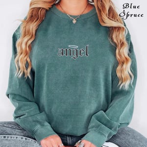 Pode incluir: Uma camisa de moletom verde com um gr&aacute;fico preto da palavra "angel" com uma aur&eacute;ola acima.