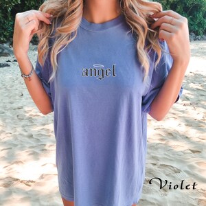 May include: A light blue t-shirt with the word "angel" printed in black with a halo above the word. The t-shirt is oversized and has a relaxed fit.