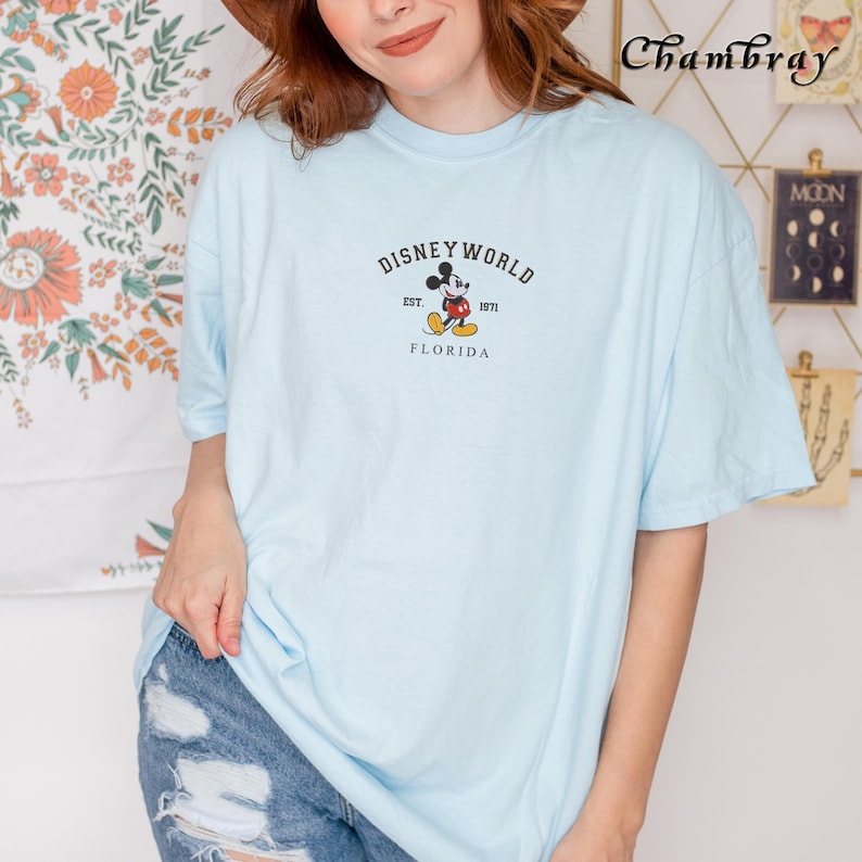 Puede incluir: Camiseta azul claro con el texto "DISNEY WORLD EST. 1971 FLORIDA" y un dibujo de Mickey Mouse. La camiseta tiene mangas cortas y cuello redondo. La camiseta est&aacute; hecha de un material suave.