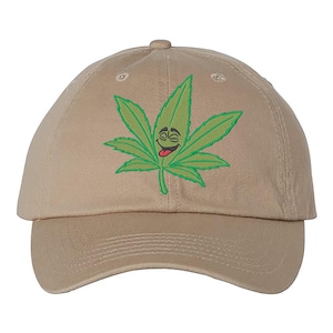 May include: A tan baseball cap featuring a green cannabis leaf with a smiling face and a red tongue. The leaf is embroidered on the front of the cap, which has a curved brim and an adjustable strap.