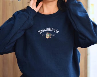 May include: Navy blue sweatshirt with the text "DisneyWorld" in a white script font. The sweatshirt features a Mickey Mouse graphic with a Santa hat and the text "EST. 1971". The word "Navy" is in the upper left corner.