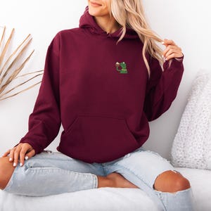 May include: A burgundy hoodie with a kangaroo pocket and a drawstring hood. The hoodie features a small embroidered design of a green frog holding a beverage. The model is wearing light blue jeans.