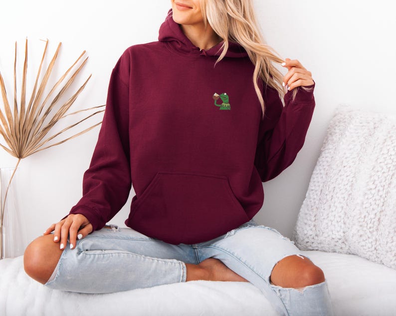 May include: A burgundy hoodie with a kangaroo pocket and a drawstring hood. The hoodie features a small embroidered design of a green frog holding a beverage. The model is wearing light blue jeans.
