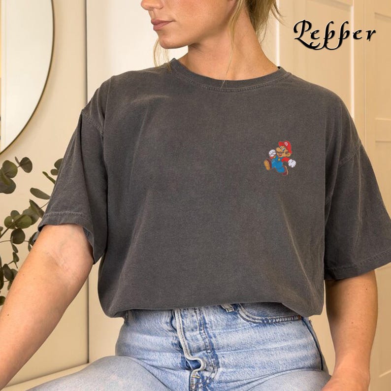 May include: A dark gray t-shirt with a small embroidered image of a video game character. The character is wearing a red hat and blue overalls. The t-shirt is worn by a person with long brown hair and is paired with light blue jeans.