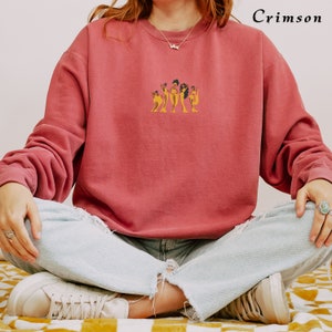 Op de afbeelding: Een crimson sweatshirt met een afbeelding van vijf vrouwen in gele jurken. De vrouwen staan in een rij en hebben donker haar.