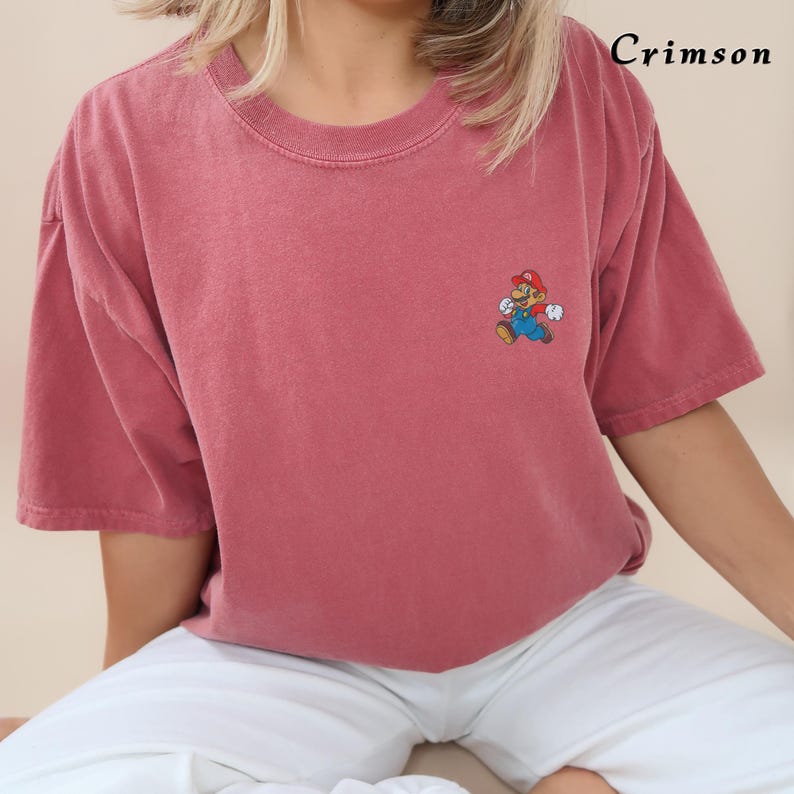 May include: A crimson red t-shirt with a cartoon character graphic of Mario from the Super Mario Bros. video game series. The character is embroidered on the shirt.