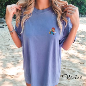 May include: A light purple t-shirt with a small embroidered image of Mario, a video game character, on the left chest. The text "Violet" is visible in the bottom right corner of the image.