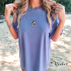 Puede incluir: Una camiseta violeta con la inscripci&oacute;n "DISNEY WORLD" y un dibujo de Mickey Mouse. Debajo del dibujo, el texto dice "EST. 1971 FLORIDA". La camiseta tiene mangas cortas y un corte holgado. La palabra "Violet" est&aacute; escrita abajo.