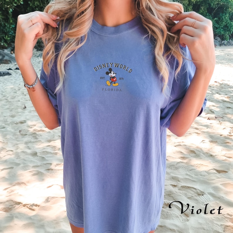 Puede incluir: Una camiseta violeta con la inscripci&oacute;n "DISNEY WORLD" y un dibujo de Mickey Mouse. Debajo del dibujo, el texto dice "EST. 1971 FLORIDA". La camiseta tiene mangas cortas y un corte holgado. La palabra "Violet" est&aacute; escrita abajo.