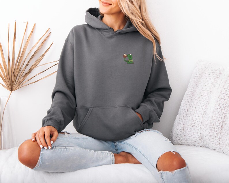 May include: A charcoal gray hoodie with a kangaroo pocket and a drawstring hood. The hoodie features a small embroidered design of a green frog holding a drink. The model is wearing light blue jeans.