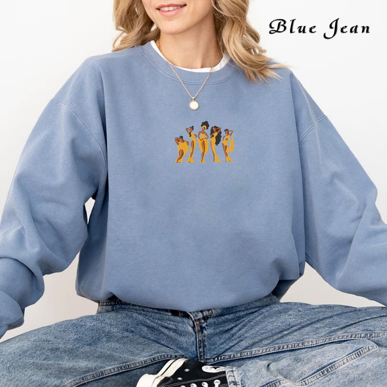 Op de afbeelding: Een lichtblauw sweatshirt met een afbeelding van vijf vrouwen in gele jurken. De tekst "Blue Jean" staat in de rechterbovenhoek van de afbeelding.